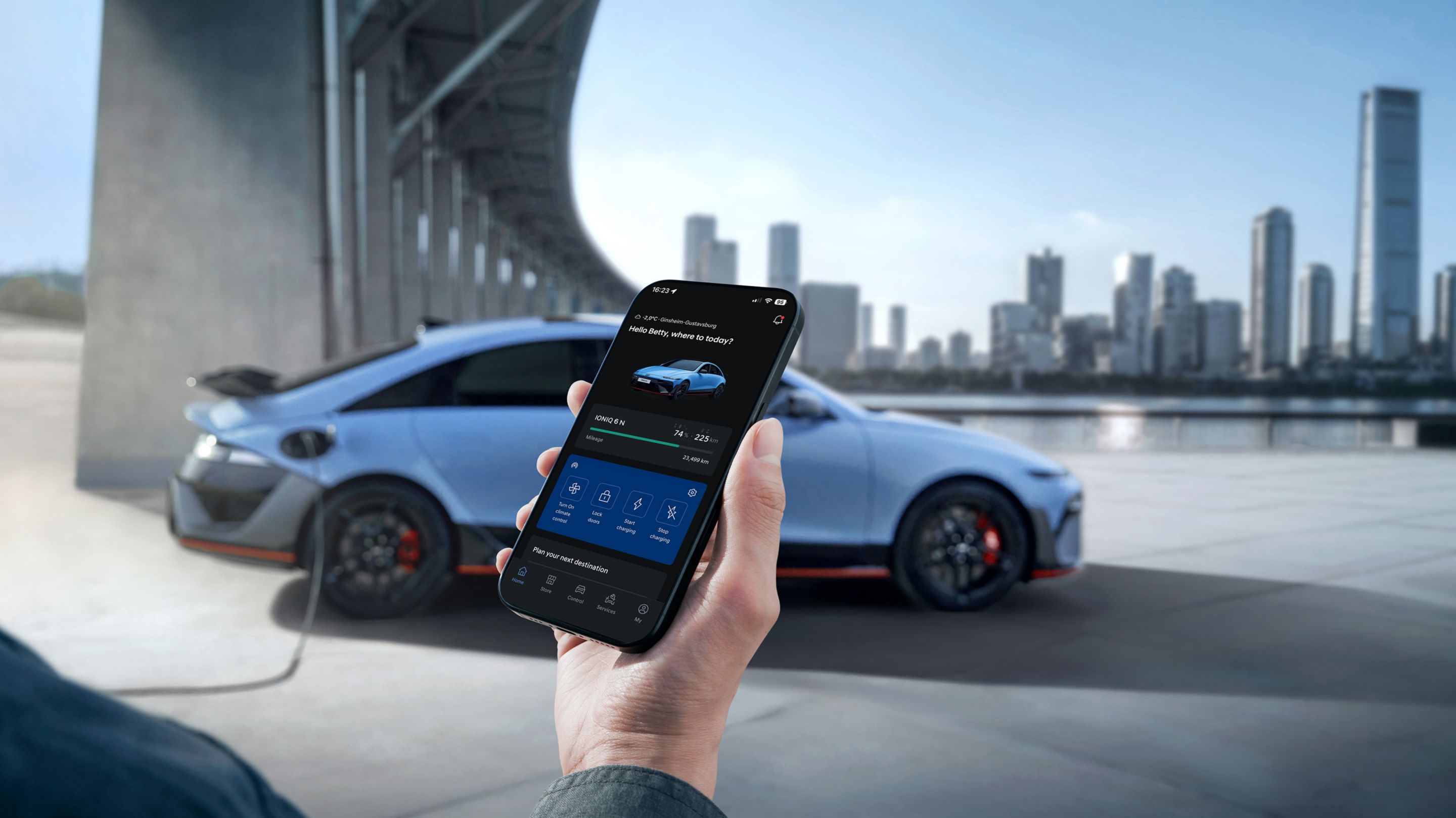 Egy kézben tartott okostelefon kijelzőjén a Hyundai IONIQ 6 N töltési státusza jelenik meg, az autóval és városi látképpel.